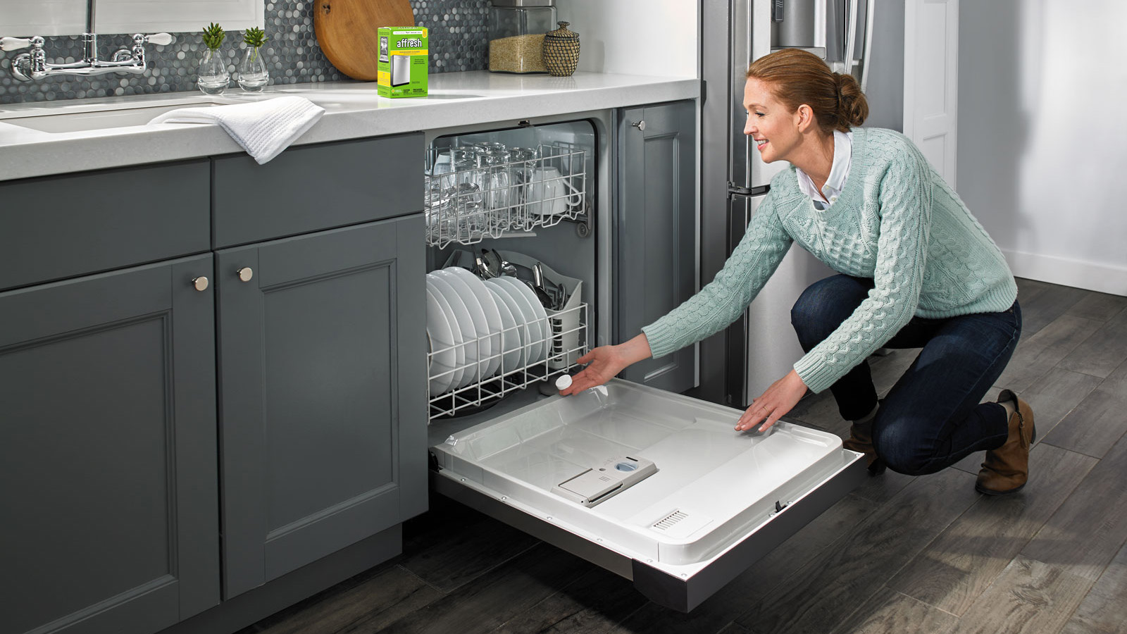 A woman crouches to pull out the bottom rack of a dishwasher. The dishwasher is filled with plates, cutlery, and glasses. On the counter above her are various items including a box of affresh® dishwasher tablets.
