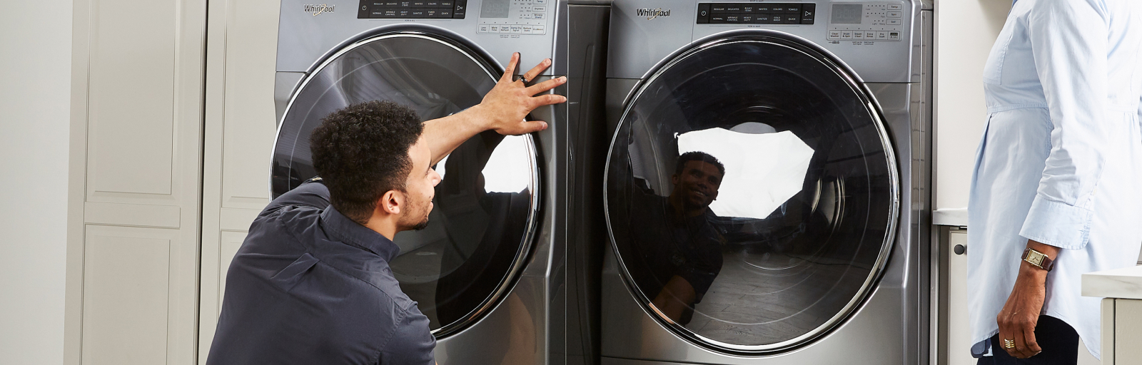 A repairman moves a laundry appliance into place next to another while someone stands next to them.