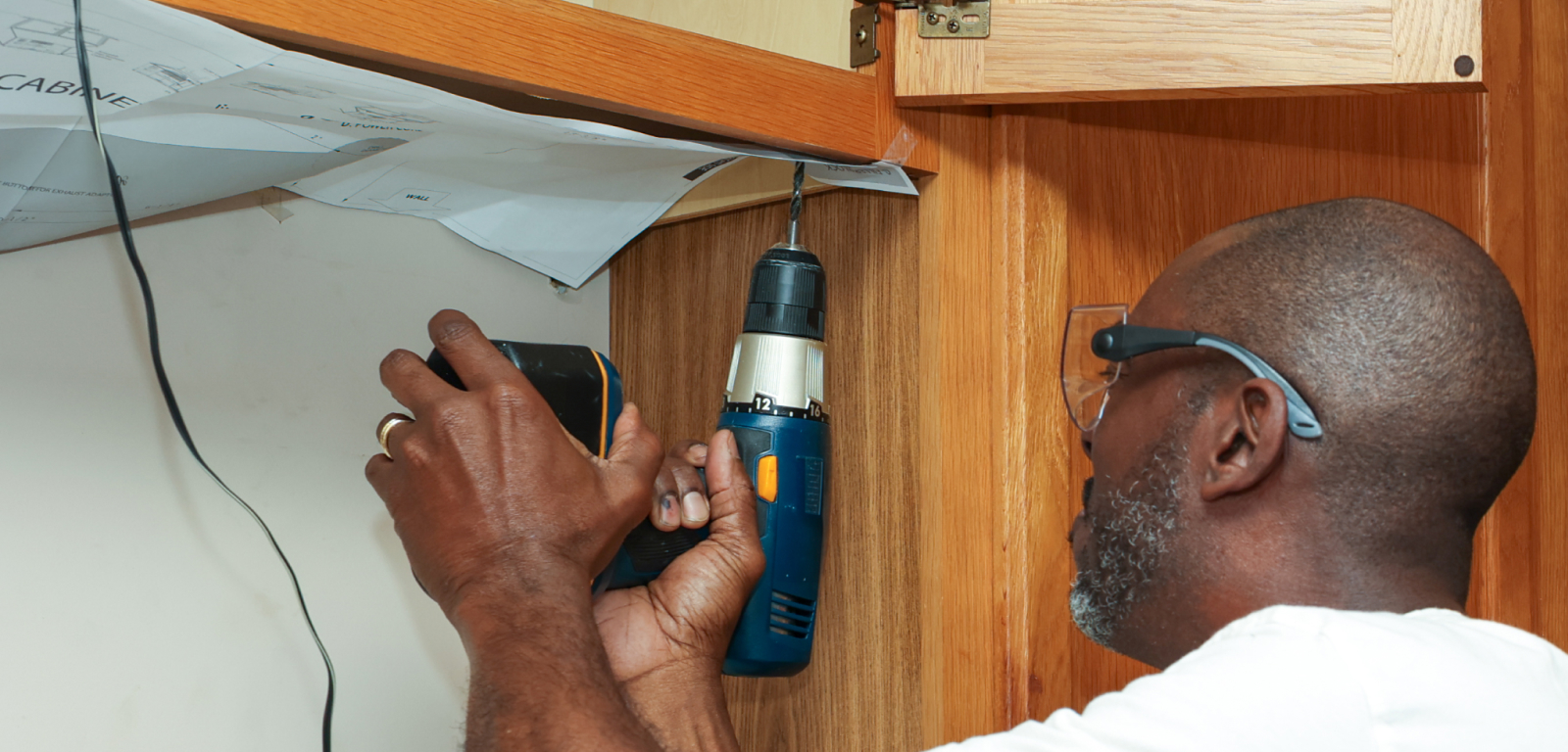 A man in safety goggles drills into cabinets from the bottom.