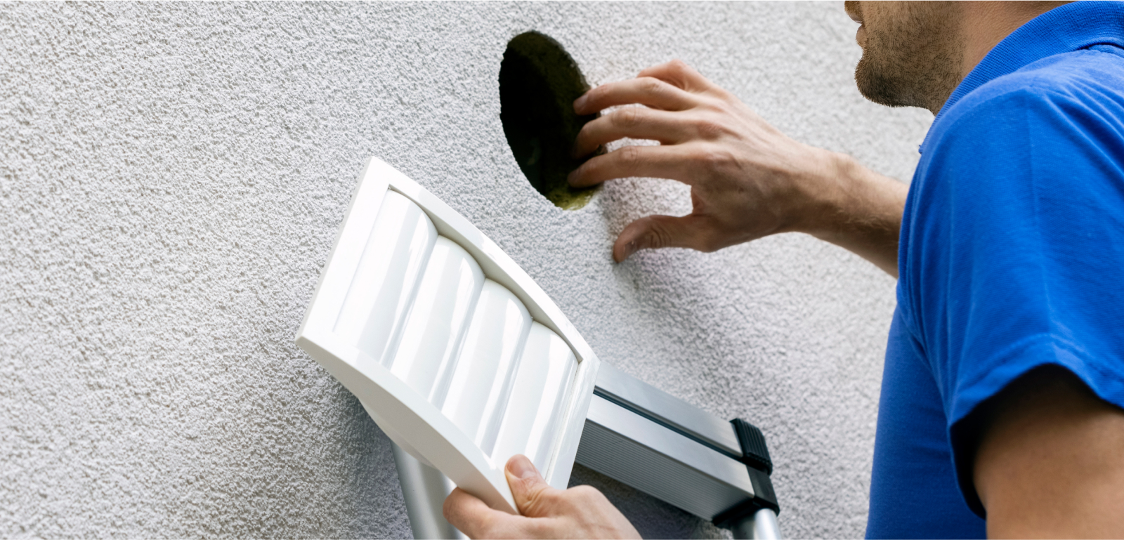 A man, holding a dryer vent cover, inspects the hole in the house that leads to the dryer.
