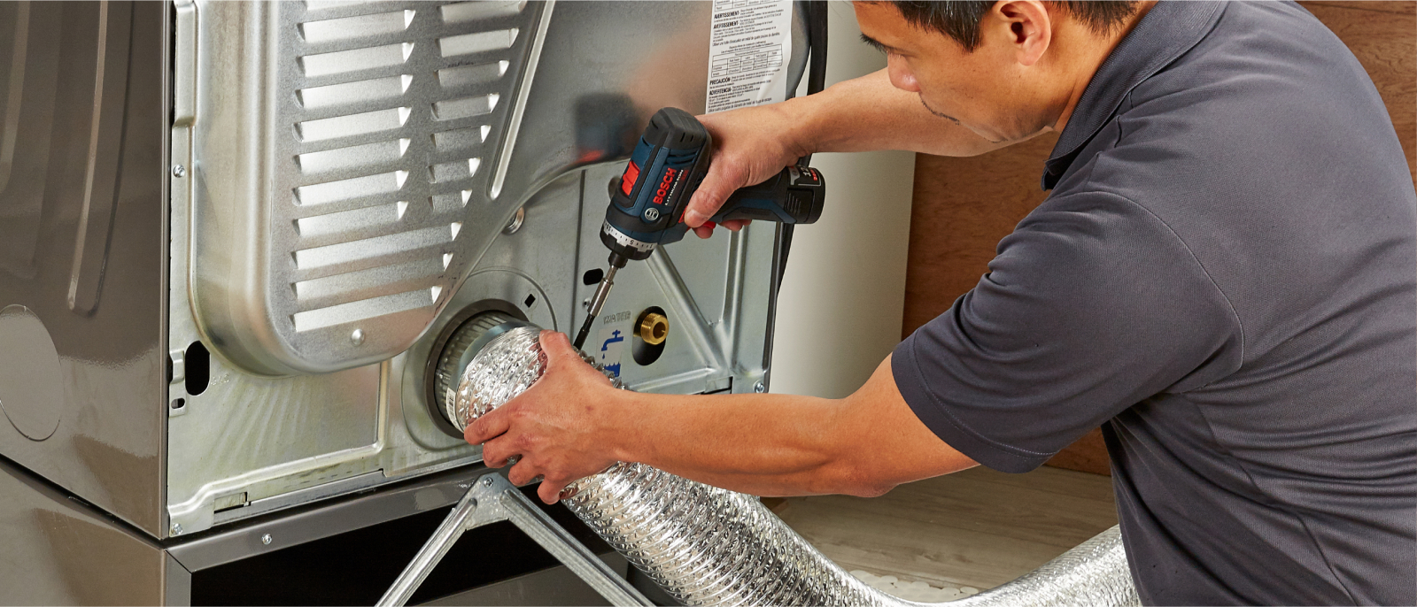 At the back of a dryer, a man drills into the dryer vent hose.