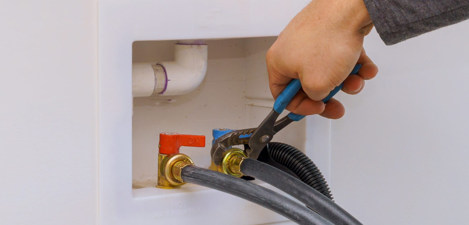 Someone uses a wrench to tighten the connection on a cold water supply on a washer. Next to it is the valve for the hot water supply.