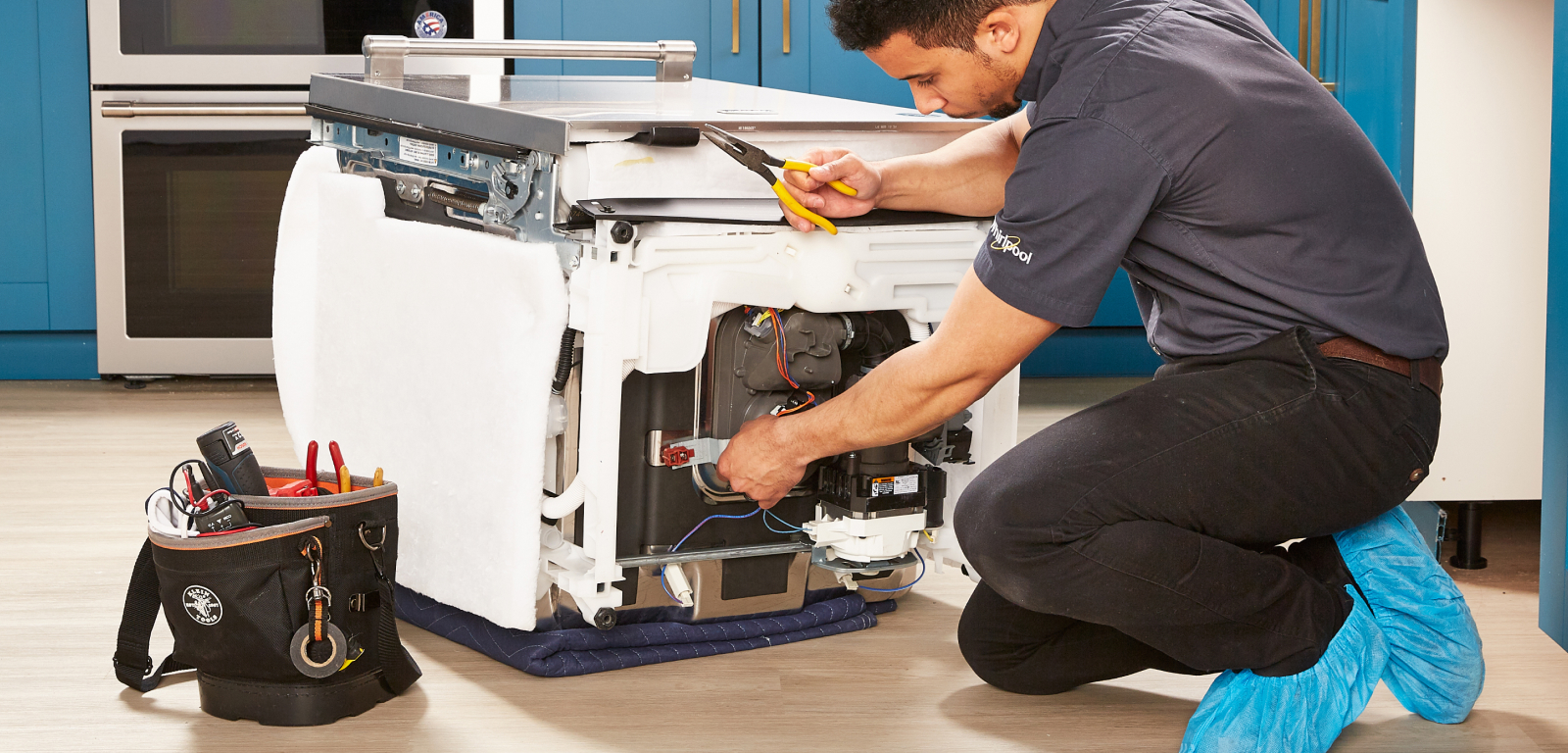 Img_Step6_1600x768 A repairman fixes a Dishwasher that's removed and on the floor. He's holding tools like a wrench and pliers. His toolbelt is next to the dishwasher.
